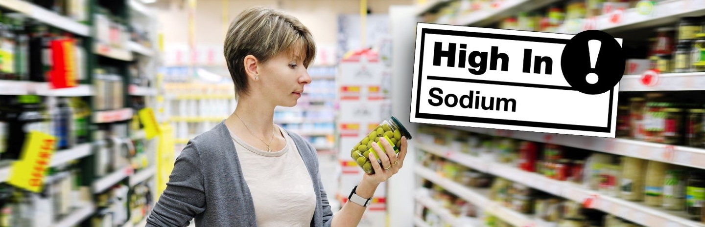 Front-of-package nutrition labeling - woman reads label on jar of olives in grocery store with High In sodium warning to the right