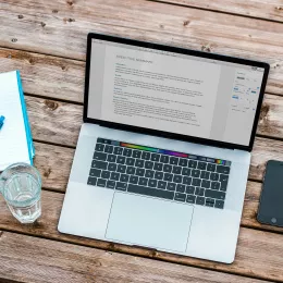 A laptop with an executive summary for a paper on the screen. A glass of water and a notepad sit beside it on a wooden table.