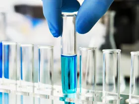 row test tubes one being held by a hand with gloves contains blue liquid