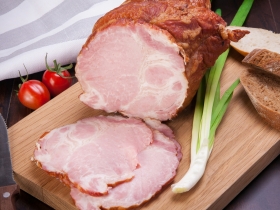 Sliced ham on a wooden cutting board beside two tomatoes, a knife, a scallion, and rustic brown bread.