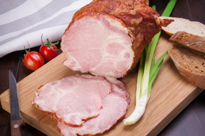 Sliced ham on a wooden cutting board beside two tomatoes, a knife, a scallion, and rustic brown bread.