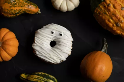 A donut decorated like a ghost surrounded by pumpkins with a black background.