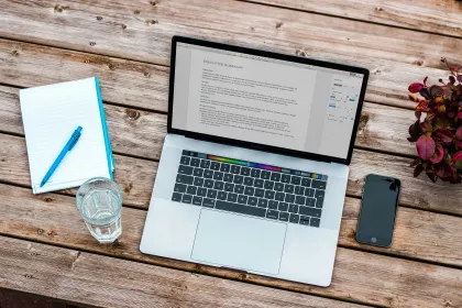 A laptop with an executive summary for a paper on the screen. A glass of water and a notepad sit beside it on a wooden table.