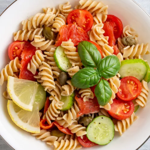 bowl of rotini with cucumber, tomatoes, olives, basil and lemon wedges