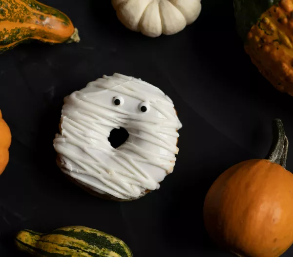 A donut decorated like a ghost surrounded by pumpkins with a black background.