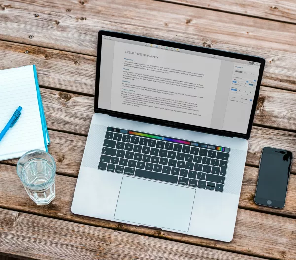 A laptop with an executive summary for a paper on the screen. A glass of water and a notepad sit beside it on a wooden table.