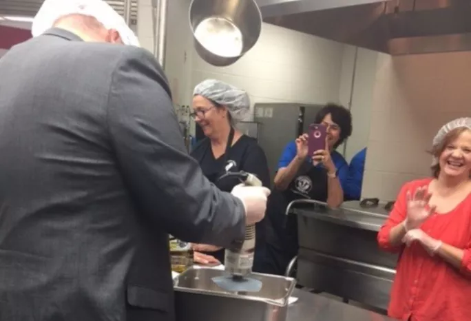 Staff in a school kitchen--several watch while one cooks