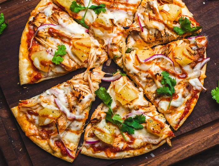 thin crust pizza on a wooden cutting board topped with pineapple, onion, parsley, chicken