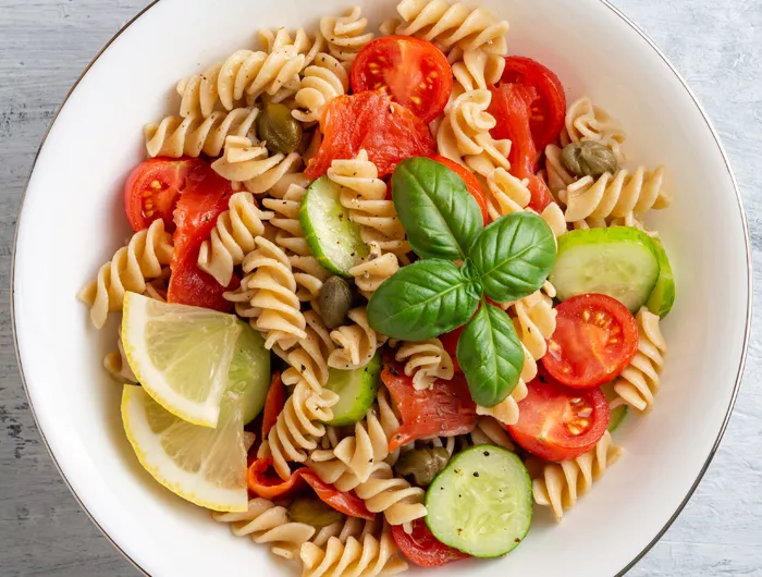 bowl of rotini with cucumber, tomatoes, olives, basil and lemon wedges