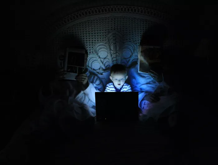 A young boy lit by the glow of an electronic screen. He is sitting between his parents, in bed, but is not being monitored.