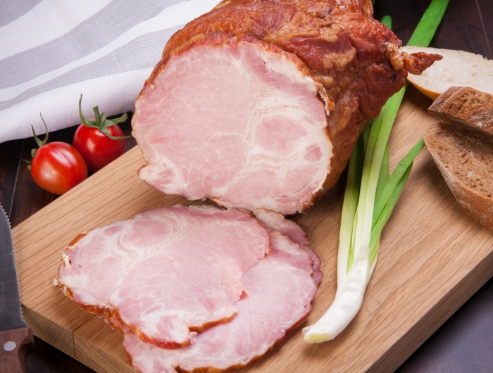 Sliced ham on a wooden cutting board beside two tomatoes, a knife, a scallion, and rustic brown bread.