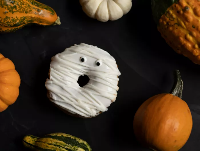 A donut decorated like a ghost surrounded by pumpkins with a black background.