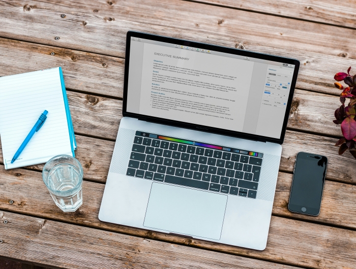 A laptop with an executive summary for a paper on the screen. A glass of water and a notepad sit beside it on a wooden table.