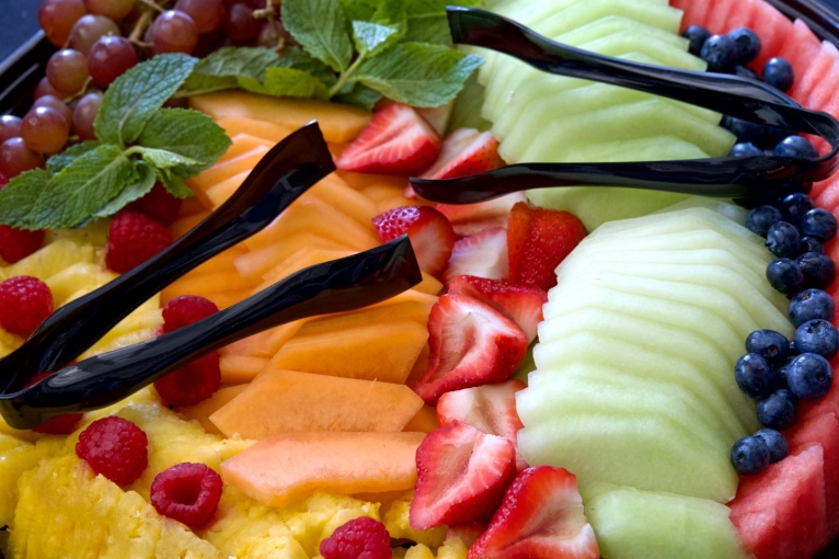 A tray of sliced fresh fruit, including strawberries, watermelon, cantaloupe, grapes and pineapple, with tongs for serving