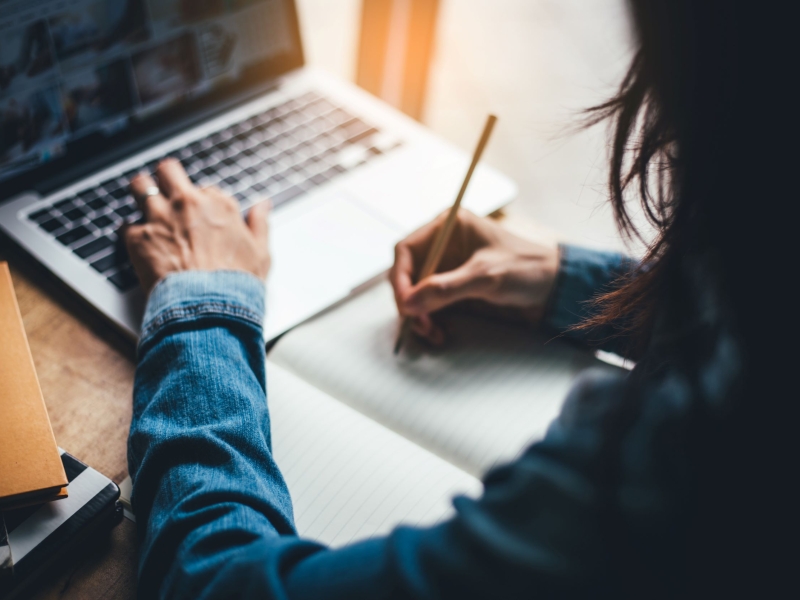 Woman writes at laptop with notebook for notes